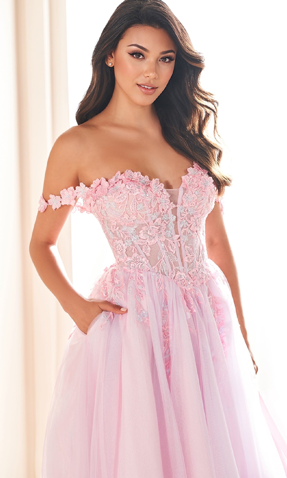 A woman wears the Ellie Wilde EW36214 long prom dress, featuring an off-the-shoulder light pink tulle a-line skirt and floral lace applique corset bodice, as she stands indoors looking at the camera.